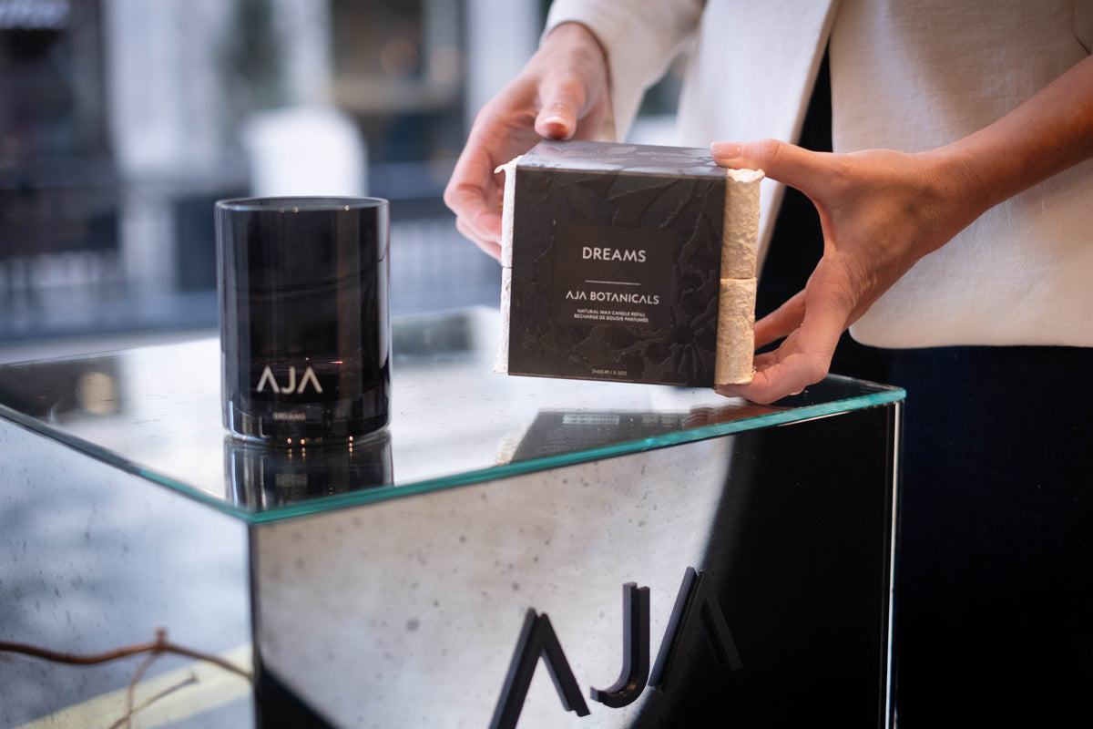 Hands holding a Dreams natural wax candle refill from Aja Botanicals beside a black Aja Botanicals candle glass